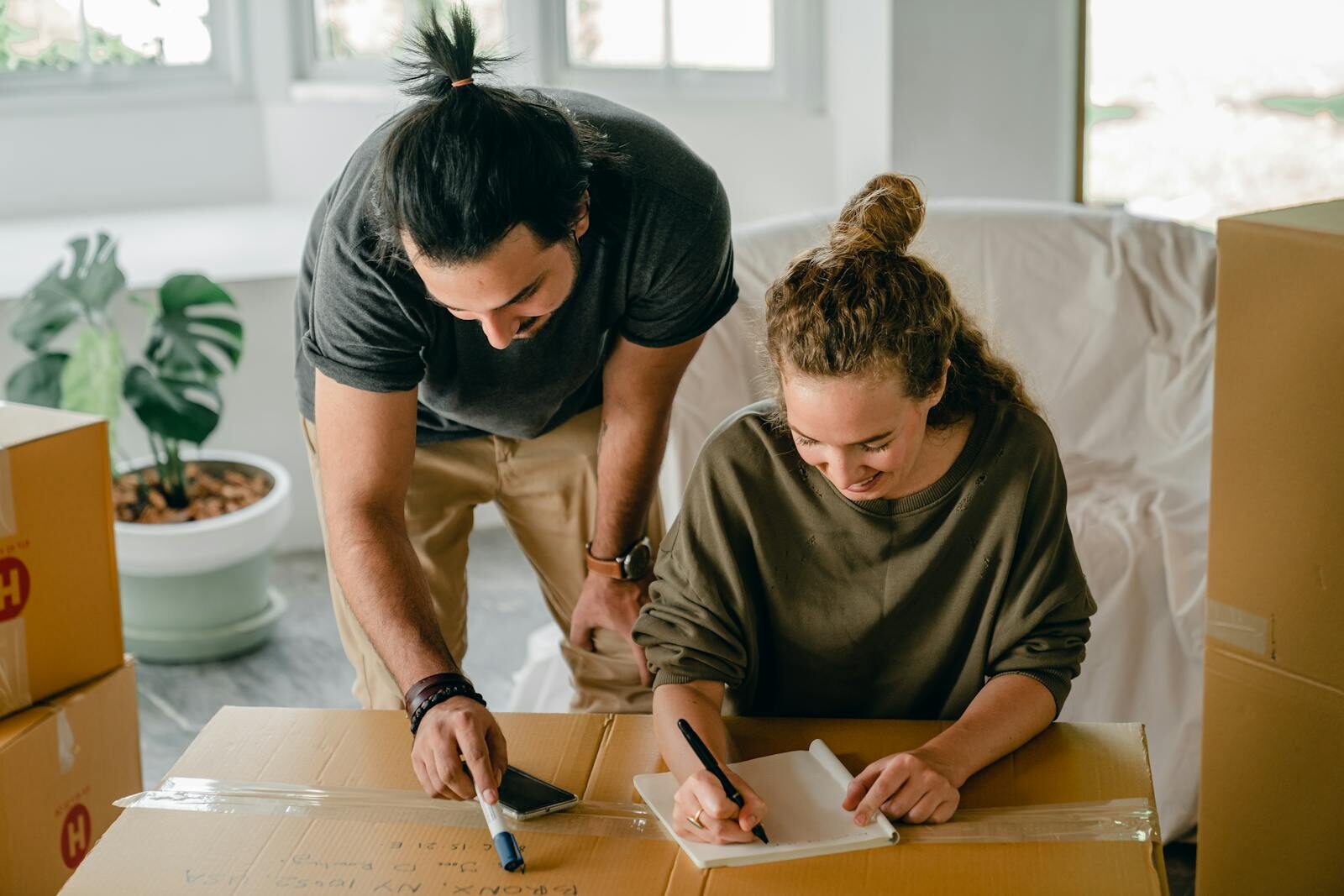 From above of ethnic man with ponytail indicating with marker at inscription on box while happy girlfriend writing with pen in notepad sitting near pile of cardboard boxes at home